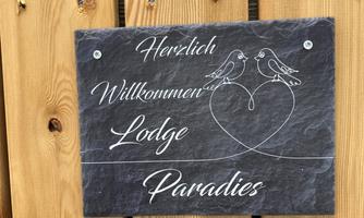 A welcome sign made of stone with the inscription "Warm Welcome Lodge Paradise" and two birds in a heart. The sign is attached to a wooden wall.