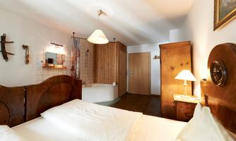 A cozy bedroom with a large double bed and rustic wooden furniture. In the background, there is a bathtub and a bright wardrobe.