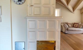 A white tiled stove stands against a wall in a bright room. In the background, furniture and a clock are visible.