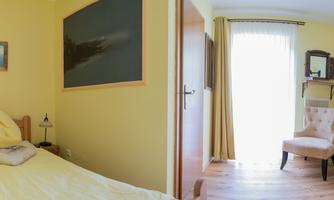 A bright room with yellow walls and a single bed. In the background, there is a chair, a mirror, and a television.