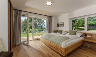 A modern bedroom with two beds and large windows. The room offers a view of the green landscape outside.