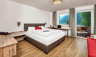 A modern bedroom with a double bed and a wooden desk. Large windows offer a view of nature.