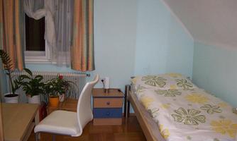 A simple single room with a bed, a desk, and a chair. There are windows with curtains and a plant in the room.