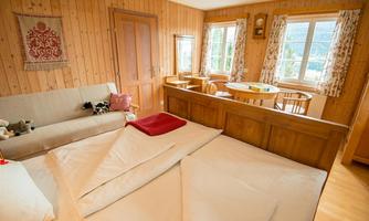 A cozy bedroom with wooden paneling, a large bed, and a sofa. Many windows let in plenty of daylight.