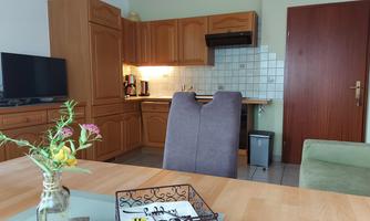 A cozy kitchen with wood-colored cabinets and a dining table. In the background, a TV and a green couch are visible.
