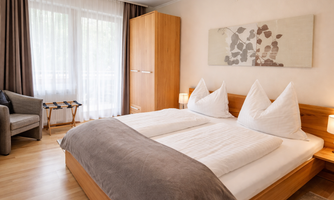 A modern bedroom with a double bed and two pillow shams. There is wooden furniture, large windows, and cozy lighting.