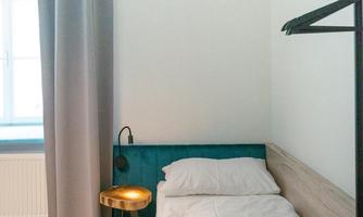 A simple bedroom with a single bed, a bedside table, and a chair. The room has a large window with a curtain and offers a calm atmosphere.