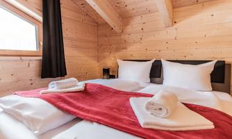A cozy bedroom with wooden walls and a large window. The beds are neatly decorated with white pillows and a red blanket.