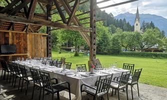 A large table with chairs is under a wooden roof in a beautiful garden. In the background, you can see a church and mountains.