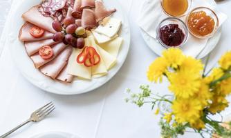 A plate with various meat products, types of cheese, and grapes. Next to it are small bowls with jam and a blooming yellow plant.