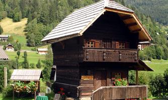 A traditional wooden house with a sloping roof and a balcony.  
Surrounded by green forests and meadows.
