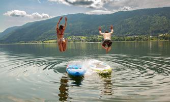 Two people are happily jumping into the water from a stand-up paddleboard. In the background, green hills and a blue sky can be seen.