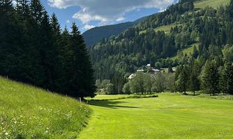 A green landscape with a wide, well-maintained path. In the background, there are mountains and a blue sky with some clouds.