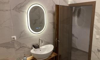 A modern bathroom with an oval mirror and an elegant sink. The walls are designed with stylish tiles, and a wooden shelf complements the high-quality look.