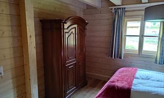 A cozy wooden room with a large, dark wardrobe and a bed with a red bedspread. The walls are made of light wood and there is a window with curtains.