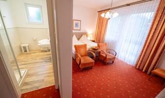 A cozy hotel room with two armchairs, a table, and a large window. In the background, the bathroom can be seen.