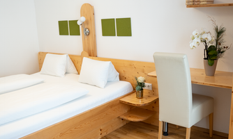A cozy bedroom with a wooden bed and two pillows. A desk with a chair and a plant completes the room.