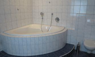 A modern bathroom with a spacious corner bathtub and white tiles. On the wall, two faucets and a toilet are visible.