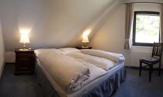 A cozy bedroom with a double bed, two nightstands, and a lamp on each side. A chair stands next to the window, letting in daylight.