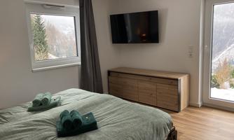 A modern bedroom with a large bed and bright wood flooring. There is a window with a view to the outside and a TV on the wall.