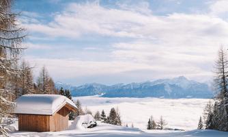 An idyllic winter landscape with a wooden house and a snow-covered area. In the background, mountains stretch under a clear blue sky.