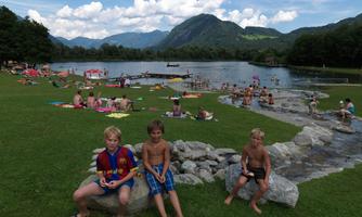 A beautiful lake with many people enjoying themselves on the shore. Children are playing and relaxing on the grass and in the water.