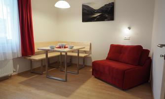 A cozy living room with a red sofa and a wooden dining area. The windows let in plenty of daylight and there is a landscape painting on the wall.