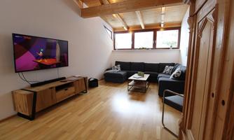 A modern living room with a large television and a cozy sofa. The room has wooden beams and large windows that let in plenty of light.