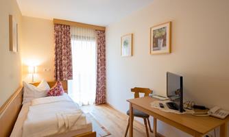 A cozy single room with a bed, a desk, and a television. The windows are decorated with curtains and the room has a warm, inviting atmosphere.