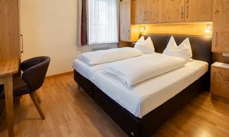 A cozy room with a double bed and wood-colored furniture. The space is bright and inviting with a desk and windows that let in natural light.