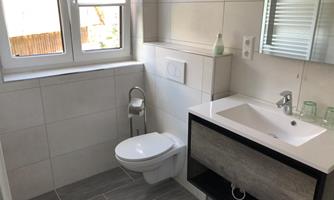 A modern bathroom with light tiles and a large window. The furnishings include a sink with a drawer and a toilet.