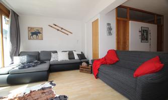 A modern furnished living room with a gray sofa and a red cushion. Skis hang on the wall and there is plenty of daylight.
