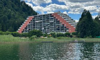 A modern building on the shore of a body of water, surrounded by trees and green landscape. The sky is partly cloudy and is reflected in the water.