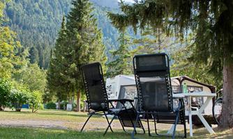 A quiet campsite with two loungers and a table outdoors. Surrounded by tall trees and mountains in the background.