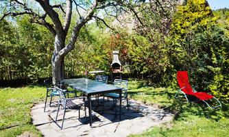 A beautiful garden table with chairs is under a tree. Next to the table is a red lounge chair and a grill.