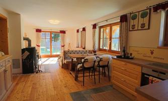 A bright and cozy kitchen with wooden flooring and large windows. In the room, there is a dining table and chairs, as well as a seating area with cozy blankets.