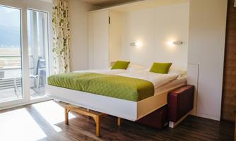 A modern bedroom with a double bed and fresh green bedding. Large windows provide plenty of daylight and a view outside.