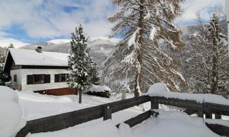 A picturesque winter house surrounded by snow-covered trees. The sky is partly cloudy, and the landscape looks peaceful and inviting.