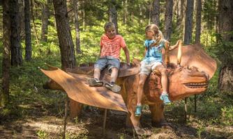 Two children are sitting on an intricately carved wooden figure in a forest. The surroundings are green and peaceful.