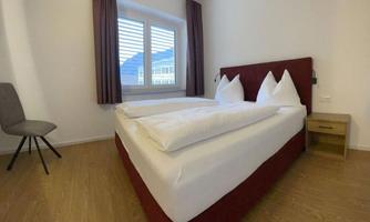 A modern hotel room with a double bed and two pillows. The walls are light, and there is a window with a view outside.