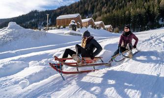 Two people are riding on a sled down a snowy slope. In the background, wooden buildings and trees are visible.