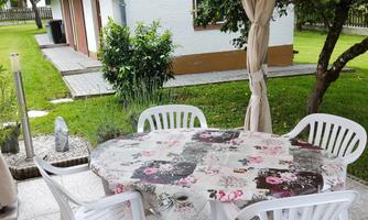 A cozy outdoor area with a round table and white chairs. In the background, a green garden and a house can be seen.