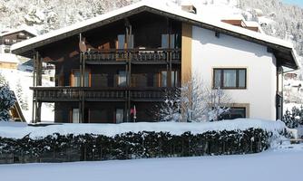 A cozy house in the snow with balconies and windows. The landscape is surrounded by snow-covered mountains.