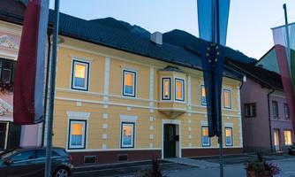 A yellow building with shining windows at dusk. In the background, mountains and flags are visible.