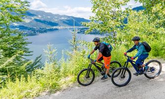 Two cyclists are riding on a forest path overlooking a lake. Surrounded by green trees and mountains in the background.