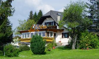An inviting house with a large balcony, surrounded by lush greenery. In the background, there are trees and a blue sky.