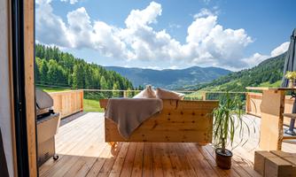 A cozy terrace with a wooden bench and a view of green mountains. The sky is blue with some clouds.