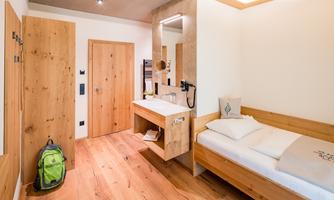 A simple room with wooden furnishings and a single bed. There is a sink, a desk, and a green bag in the corner.