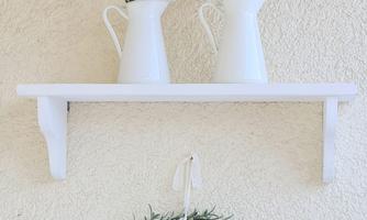 A white wall with a shelf on which are two vases with flowers. Below hangs a round wreath made of green leaves.