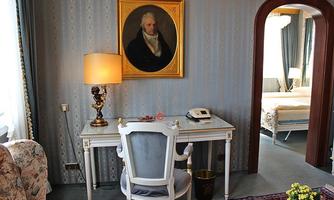An elegant room with a desk, a chair, and an old painting on the wall. In the background, there is a bed and a cozy seating area.